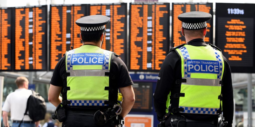 Two British Transport Police officers, seen from behind, stand in front of orange departure boards at a busy railway station.