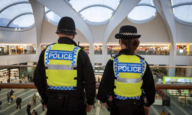 Two British Transport Police officers in high-visibility vests, seen from behind, looking out from a balcony over a busy railway station concourse.