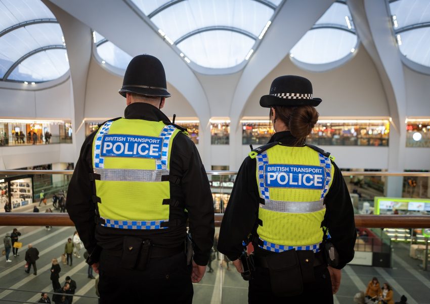 Two British Transport Police officers in high-visibility vests, seen from behind, looking out from a balcony over a busy railway station concourse.