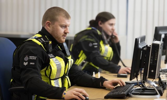 A police sergeant in a high-visibility vest types at a desktop computer in an office, while a colleague behind him makes a phone call at the next desk.