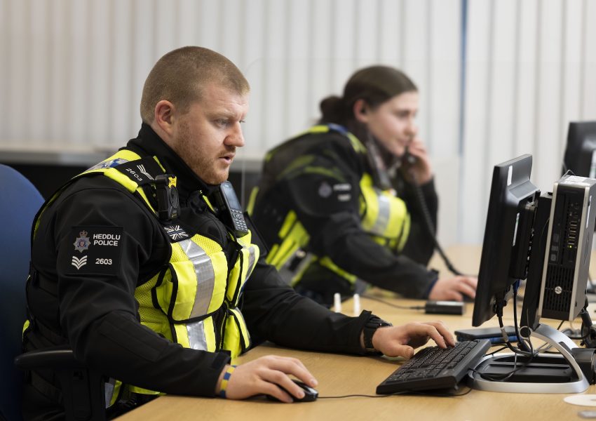 A police sergeant in a high-visibility vest types at a desktop computer in an office, while a colleague behind him makes a phone call at the next desk.